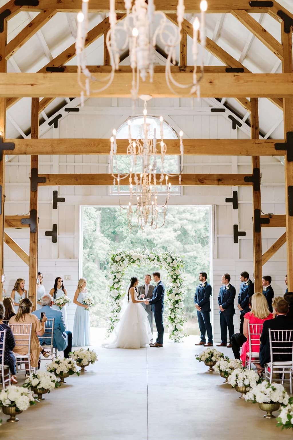 A wedding ceremony under the chandeliers in the white barn, guests seated on both sides
